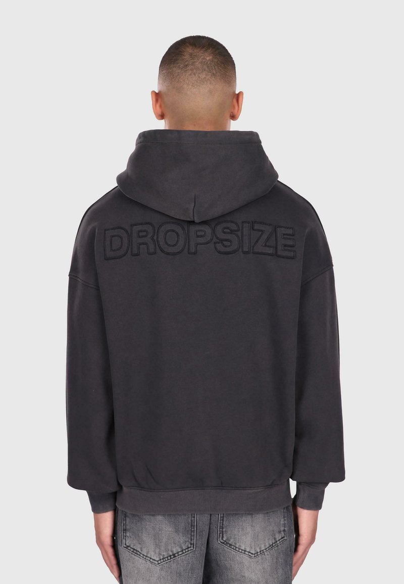 Mand med kort hår iført en mørkegrå hættetrøje med "DROPSIZE" broderet på den øverste del af ryggen og slidte grå jeans.