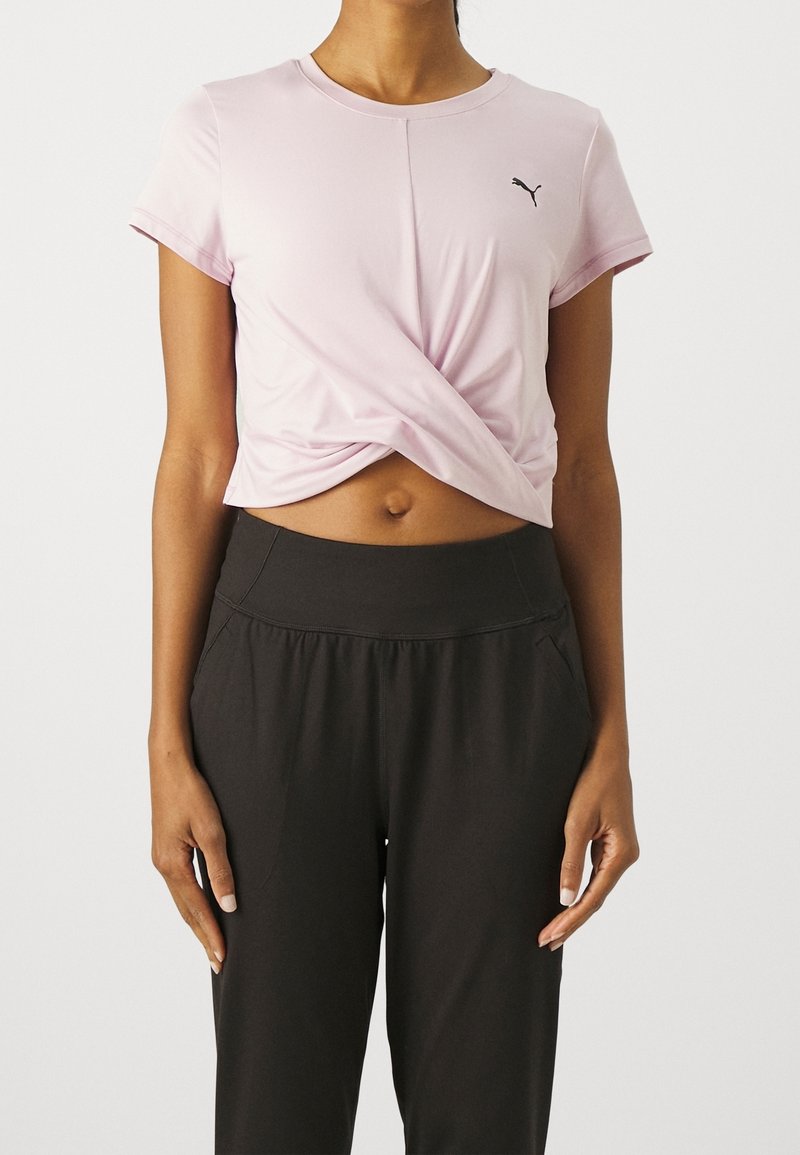 Camiseta corta de manga corta en color rosa claro con detalle de frente retorcido y pequeño logo negro. Combinada con pantalones negros de tiro alto.