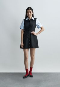 Νεαρή γυναίκα στέκεται μπροστά σε ουδέτερο φόντο, φορώντας ένα μαύρο αμάνικο φόρεμα με κουμπιά πάνω από ένα ανοιχτό μπλε κοντομάνικο πουκάμισο, κόκκινες κάλτσες και μαύρα παπούτσια.