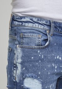Κοντινή λήψη από μπλε distressed τζιν με λευκές πιτσιλιές μπογιάς και ξεφτισμένη τρύπα, φορεμένα με λευκό ριμπ μπλουζάκι.