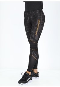 Black leggings with a geometric print and gold "SWEDEMOUNT" text, featuring a high waistband and contrasting yellow stitching details. Black sneakers worn.