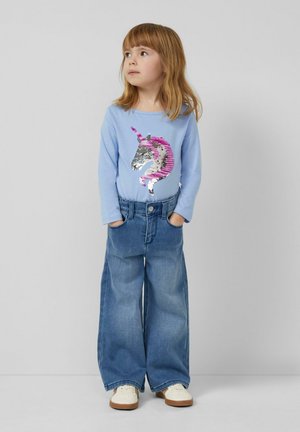 Junges Mädchen mit langen Haaren, trägt ein blaues Langarmshirt mit einem Pailletten-Einhorn, weite Jeans und weiße Schuhe, die Hände in den Taschen.