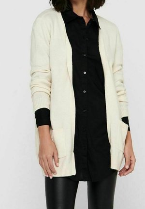 Crème kleurige cardigan van zachte stof met een open voorkant, lange mouwen en twee voorzakken, gedragen over een zwart overhemd met knopen.
