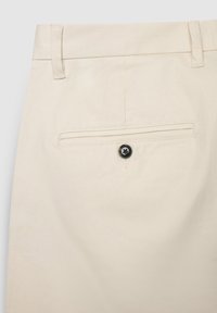 Hellbeige Hose mit schlanker Passform, ausgestattet mit einer Gesäßtasche und einem schwarzen Knopfakzent. Das Material wirkt glatt und flexibel.