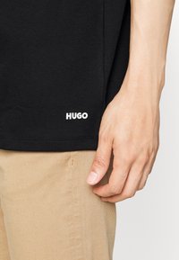 T-shirt nera Hugo con maniche corte, con un logo bianco all'orlo. Pantaloni khaki chiaro mostrati nella parte inferiore.