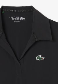 Camisa polo negra de corte regular, con un cuello sutil, logo del cocodrilo verde bordado y un cuello texturizado con el nombre de la marca.
