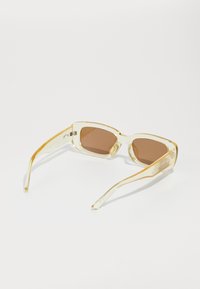 Gafas de sol transparentes amarillas con lentes marrones. Montura cuadrada, diseño elegante y brazos curvados con un sutil logotipo en la patilla.
