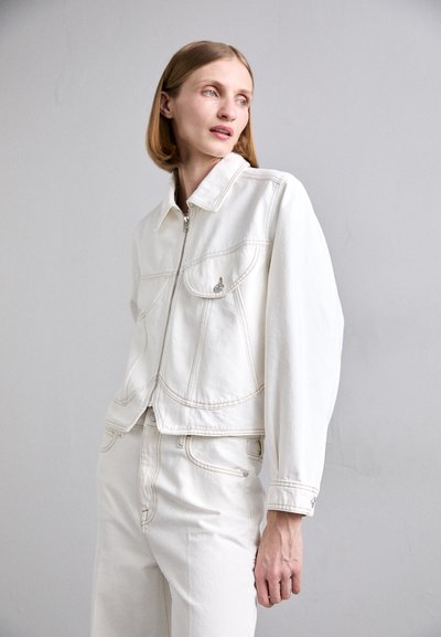 Weiße Jeansjacke mit gekürztem Schnitt, Reißverschluss, spitzem Kragen und zwei Fronttaschen. Besitzt sichtbare Nähte und metallische Akzente.