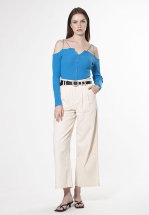 Blaue gerippte Strickjacke mit kaltem Schulterdesign, schwarzer Gürtel mit runder Schnalle, cremefarbene weit geschnittene Hose und schwarze Riemensandalen.