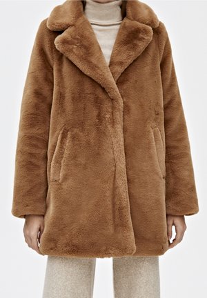 Manteau marron en fausse fourrure avec grands revers porté sur un col roulé beige et un pantalon texturé assorti, visible des épaules jusqu'à mi-cuisse.