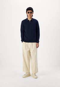 Pullover navy a maniche lunghe con colletto a mezza zip, pantaloni leggeri beige, sneakers bianche e occhiali da sole rotondi. Stile minimalista.