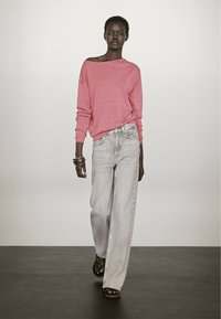 Różowy sweter hiszpański, jasnoszare szerokie dżinsy, brązowe sandały. Modelka stoi na neutralnym tle, prezentując luzacki strój.