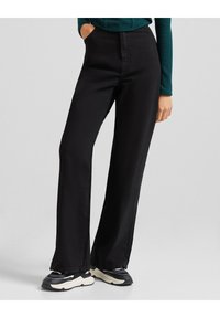 Pantalones de pierna ancha negros, hechos de una tela suave con un corte entallado, que incluyen bolsillos laterales y se combinan con zapatillas negras y blancas.