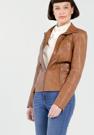 Vrouw in een bruine leren jas over een witte kanten blouse en blauwe jeans, staand tegen een eenvoudige lichte achtergrond, kijkend naar voren.