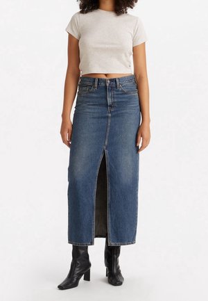 Kvinde iført en lysegrå cropped t-shirt, højtaljet lang blå denim nederdel med frontslids og sorte hælede støvler, stående mod en hvid baggrund.