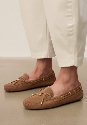 Pieds portant des mocassins beige en daim avec des détails de nœud, associés à un pantalon court de couleur crème sur un sol neutre.