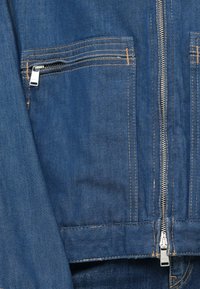 Μπλε τζάκετ από τζιν με μπροστινό φερμουάρ, δύο τσέπες με φερμουάρ και ασημένια εξαρτήματα, και αντίθετη κίτρινη ραφή. Λείο υλικό.