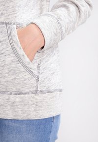 Světle šedá mikina s kapucí z texturované tkaniny, se dvěma kapsami vpředu a žebrovanými manžetami. Švy mají kontrastní stehy.