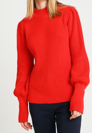Roter gerippter Pullover mit hohem Kragen, langen Ballonärmeln und einem strukturierten Strickdesign. Kombiniert mit dunklen Hosen.
