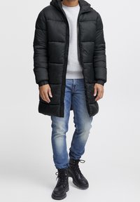 Zwarte pufferjas met doorgestikt ontwerp, capuchon, elastische manchetten en volledige rits aan de voorkant, gecombineerd met lichtblauwe jeans en zwarte veterschoenen.