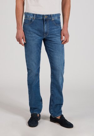 Mann trägt mittelblaue Jeans mit geradem Bein, weißes Hemd und dunkelblaue Loafers, steht vor einem einfarbigen hellen Hintergrund.