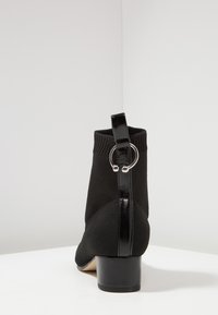 Bottine noire à cheville avec une tige en tissu tricoté, un talon brillant et élégant, et une bride décorative ornée d'un accent métallique circulaire.