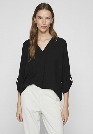 Zwarte, losse blouse met een V-hals, driekwart mouwen en opgerolde knoopaccenten, gecombineerd met witte broeken. Gestructureerde stof.