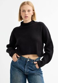 Suéter de punto negro recortado con textura acanalada y cuello alto, combinado con jeans de mezclilla azul de cintura alta. Mangas sueltas prominentes.