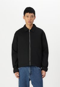 Veste zippée noire avec un col pointu, deux poches avant et une coupe décontractée. Associée à un jean en denim bleu ample.