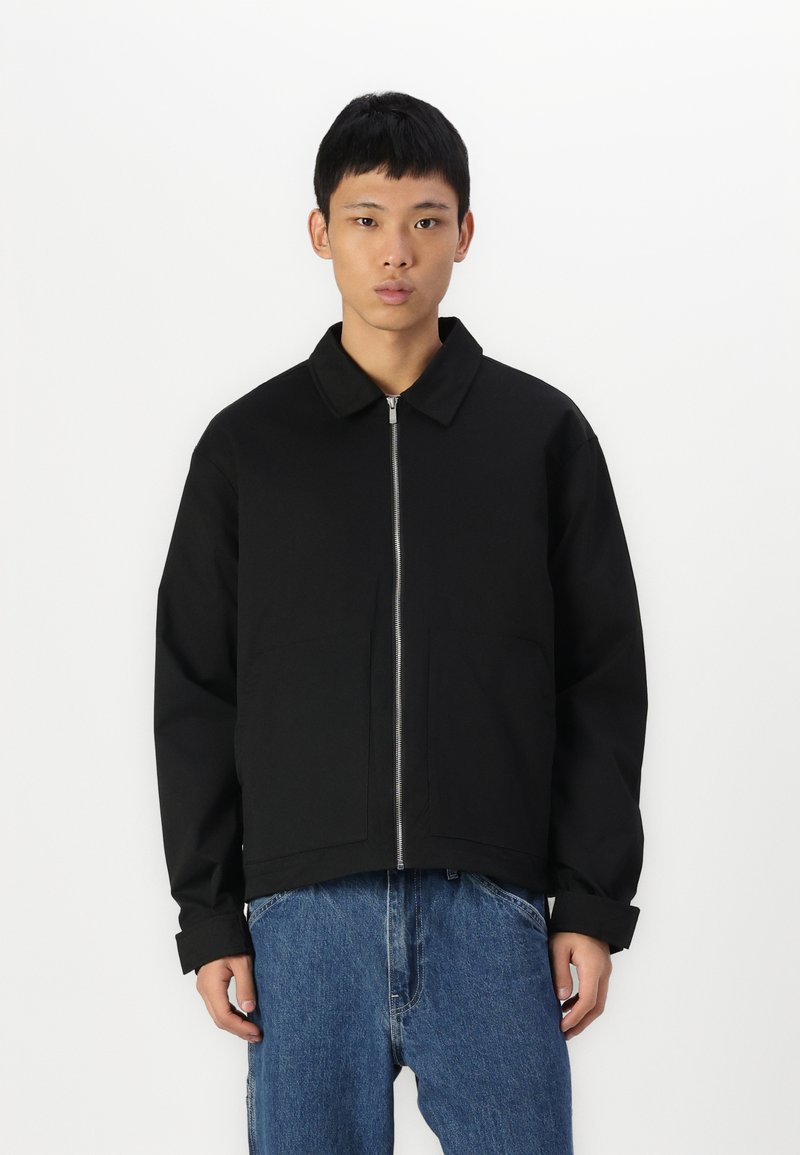 Veste zippée noire avec un col pointu, deux poches avant et une coupe décontractée. Associée à un jean en denim bleu ample.