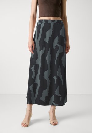 Femme portant une longue jupe noire et grise à motif abstrait, un top court marron et des sandales transparentes à talons hauts, debout devant un fond uni.