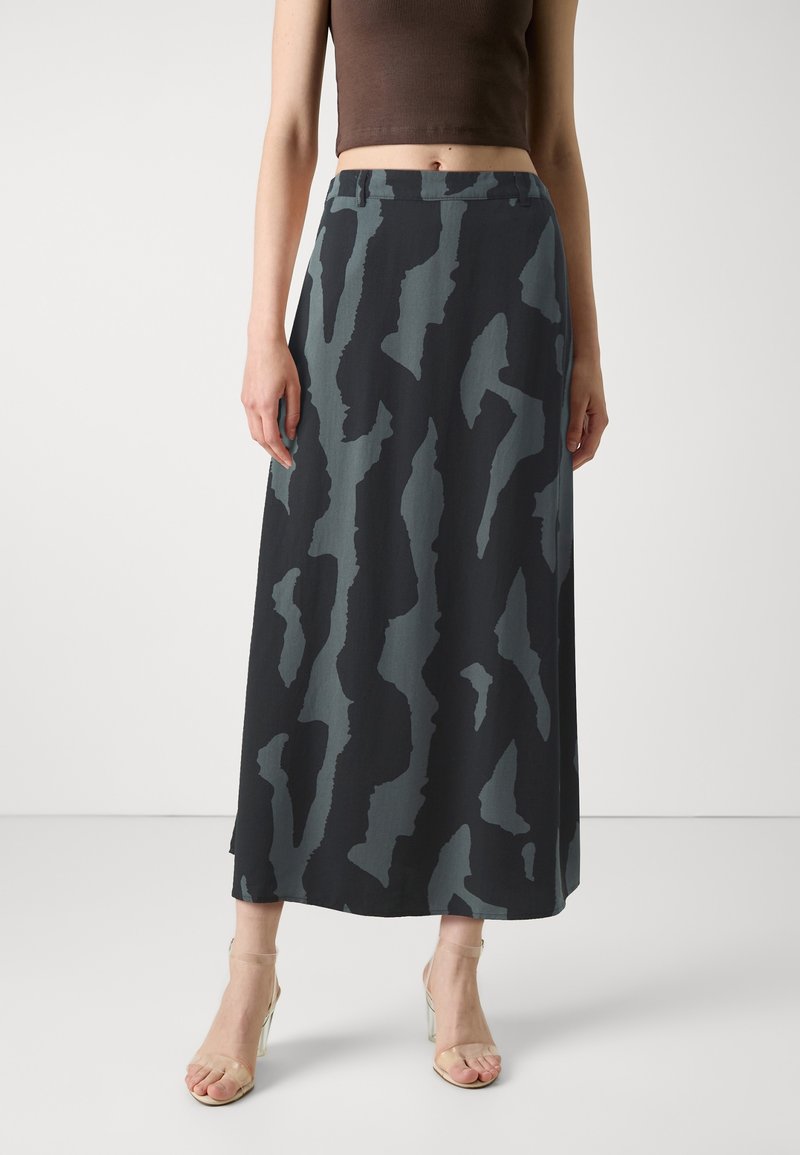 Femme portant une longue jupe noire et grise à motif abstrait, un top court marron et des sandales transparentes à talons hauts, debout devant un fond uni.
