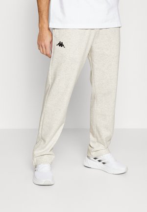 Mężczyzna ubrany w jasnobeżowe dresy z czarnym logo na lewym udzie oraz białe buty sportowe z czarnymi paskami.