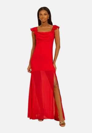 Femme portant une robe de soirée rouge sans manches avec des bretelles à volants et une jupe inférieure transparente, posant avec une jambe exposée à travers une fente latérale.