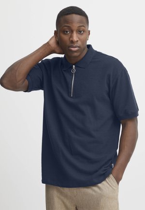 Navy short-sleeve polo shirt with a zippered collar and soft texture, paired with light-colored, textured pants.
