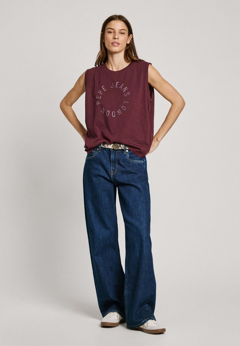 Bordeaux mouwloze t-shirt met een cirkelvormige "Pepe Jeans London" grafiek, gecombineerd met losse donkerblauwe jeans en witte sneakers.