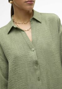 Camicia verde chiaro con bottoni in tessuto testurizzato, caratterizzata da un colletto, maniche lunghe e piccoli bottoni. Collana a catena dorata a strati visibile.