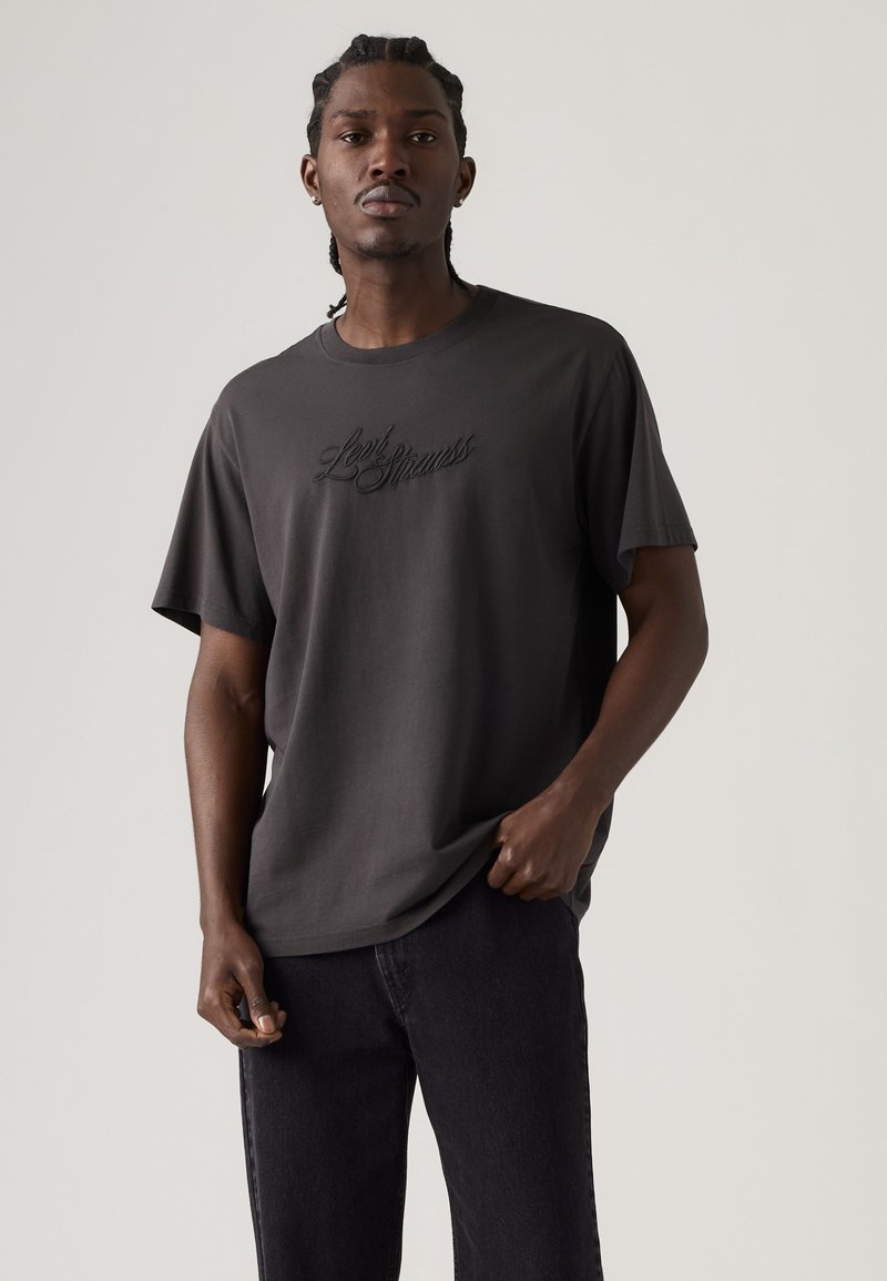 Man with braided hair wearing a dark gray Levi Strauss t-shirt and black jeans standing against a plain light background.