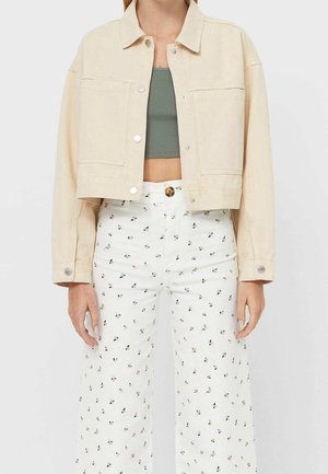 Femme portant une veste courte beige sur un crop top vert, associée à un pantalon blanc taille haute avec un petit imprimé floral.
