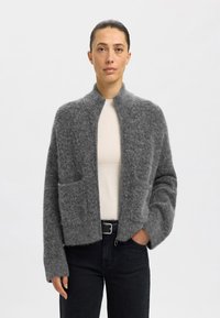 Grå stickad cardigan med en strukturerad yta, hög krage och två framfickor, kombinerad med en beige topp och mörka jeans.