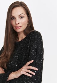 Black knitted sweater with a textured surface featuring small sequins. Loose fit with a wide neck and ribbed cuffs. Model poses with hands crossed.