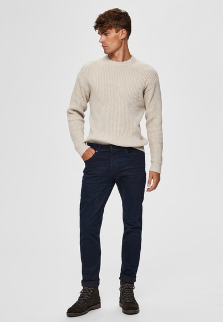 Suéter de malha canelada creme, jeans slim-fit azul-escuro e botas escuras; modelo está em fundo branco, virando-se ligeiramente para a esquerda.