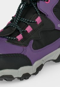 Gros plan d’une chaussure de randonnée violette et noire avec une semelle texturée et des lacets noirs à pois bleus passés dans des œillets roses.