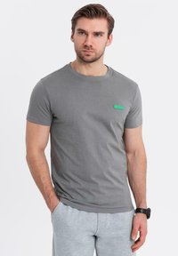 T-shirt grigia in cotone con scollatura rotonda, maniche corte e una piccola etichetta verde del marchio sul petto. Texture liscia, vestibilità casual.