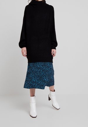 Schwarzer, gestrickter Over-size-Pullover mit hohem Kragen und langen Ärmeln, kombiniert mit einem blauen Rock mit gepunktetem Muster und weißen Knöchelstiefeln.