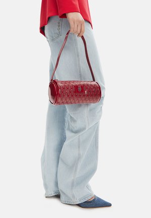 Osoba držící červenou válcovou kabelku, oblečená v modrých širokých džínách, modrých špičatých botách a červeném topu s dlouhým rukávem.