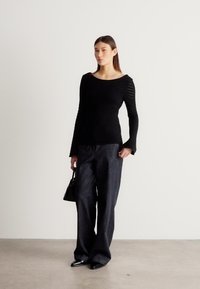 Fekete hosszú ujjú, texturált felső enyhe kivágásokkal, sötét széles szárú farmernadrággal és egy kis fekete kézitáskával párosítva.