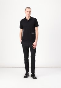 Fekete póló ing rövid ujjal, galléros nyakkal és fehér szöveges részletekkel az elején, fekete, slim-fit nadrággal és fekete cipővel párosítva.