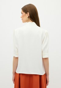 Blusa bianca con maniche corte arrotolate, texture liscia e bottoni ai polsini, abbinata a una gonna color ruggine. Vista dal retro.