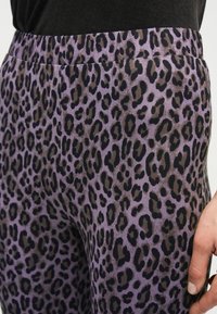 Μωβ κολάν με μαύρο και σκούρο καφέ σχέδιο λεοπάρ, φορεμένο με μαύρο τοπ, που αποκαλύπτει τη μέση και το χέρι ενός ατόμου στο πλάι.